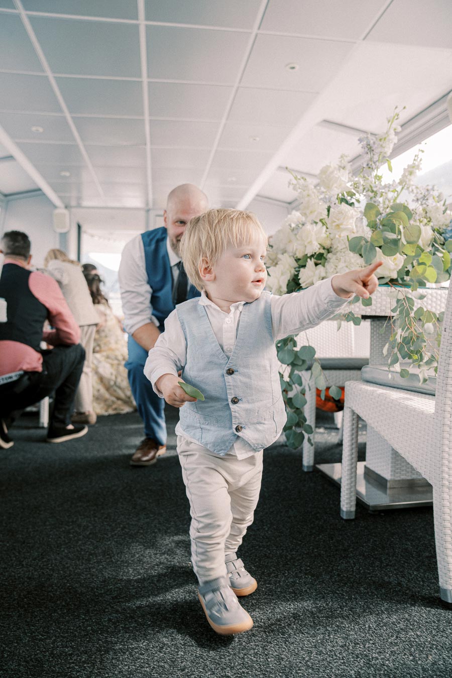 Toddler in a light blue vest pointing excitedly indoors, with adults and floral decorations in the background.