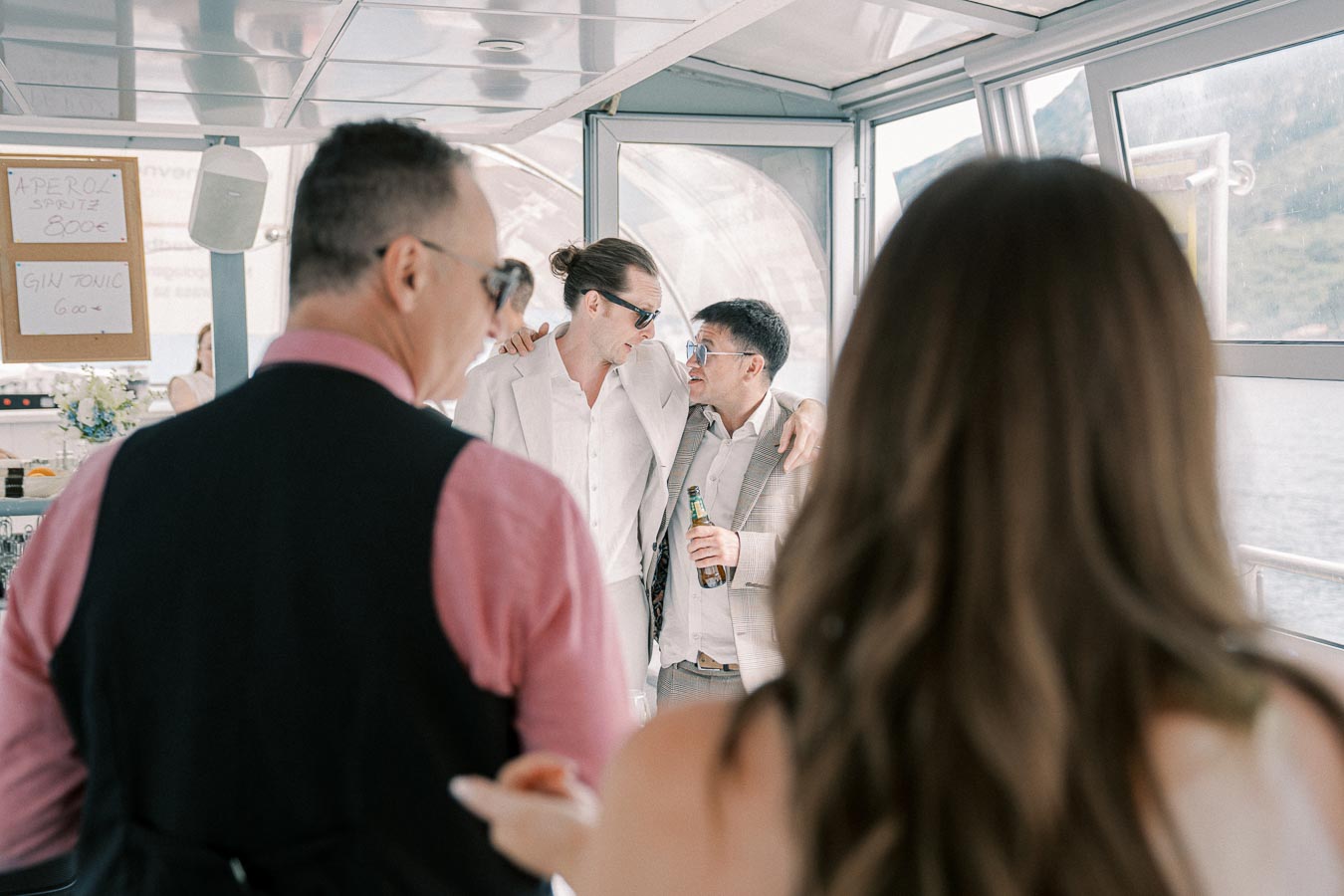 Group enjoying a celebration aboard a boat, featuring two men in formal attire hugging and conversing, with drinks in hand, surrounded by friends and a scenic water view in the background.
