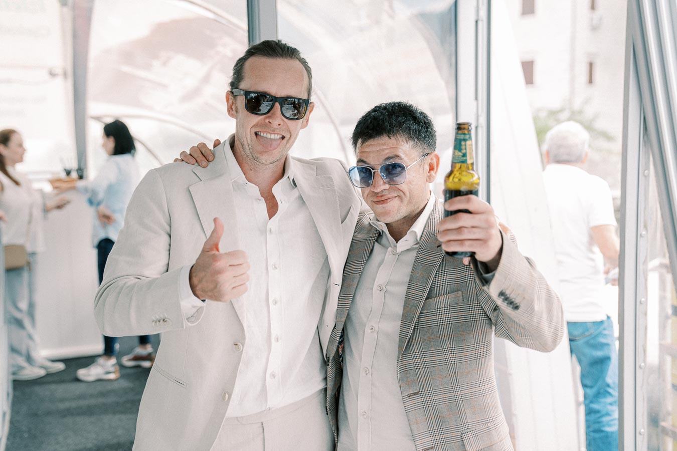 Two men in stylish suits and sunglasses enjoy a social gathering indoors, with one giving a thumbs-up and the other holding a beer.