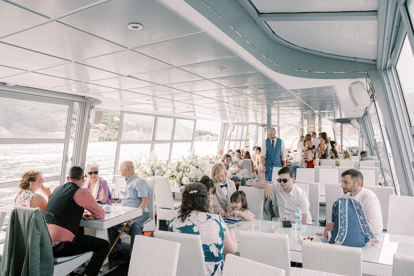 A group of people enjoying a social gathering on a scenic river cruise, seated at elegantly decorated tables with flowers, surrounded by panoramic views of the water and distant hills through large windows.