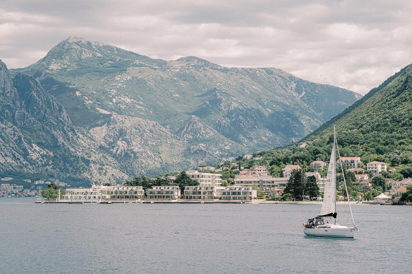 Sailboat navigating a scenic bay with mountainous landscape and waterfront buildings in the background on a cloudy day.
