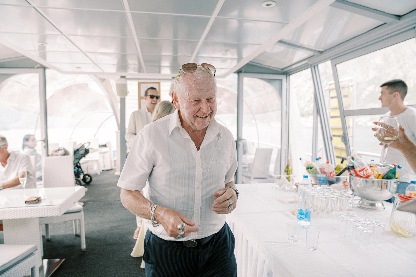 Elderly man enjoying a lively gathering on a boat, surrounded by friends and refreshments. The bright, modern interior and cheerful atmosphere suggest a celebration or party.