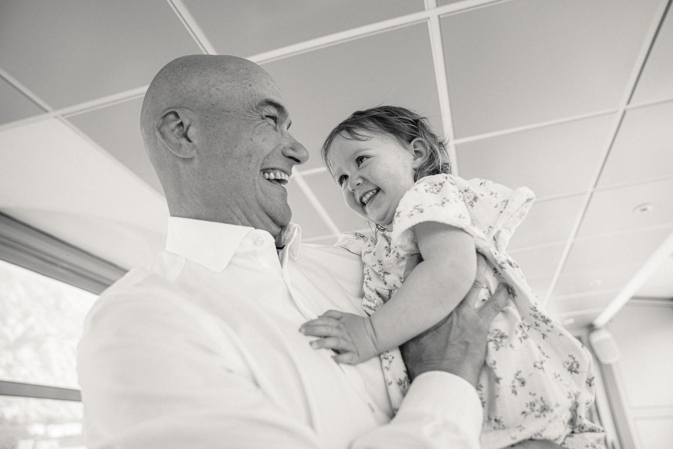 Joyful moment between an older man and a young girl, both smiling at each other, captured in black and white inside a modern building.