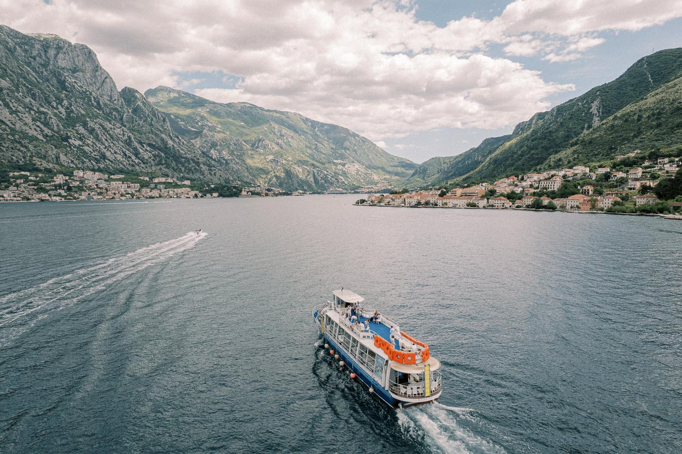 A scenic panoramic view of a boat cruising through the majestic waters of a fjord surrounded by lush mountains and a charming coastal town under a blue, partly cloudy sky.