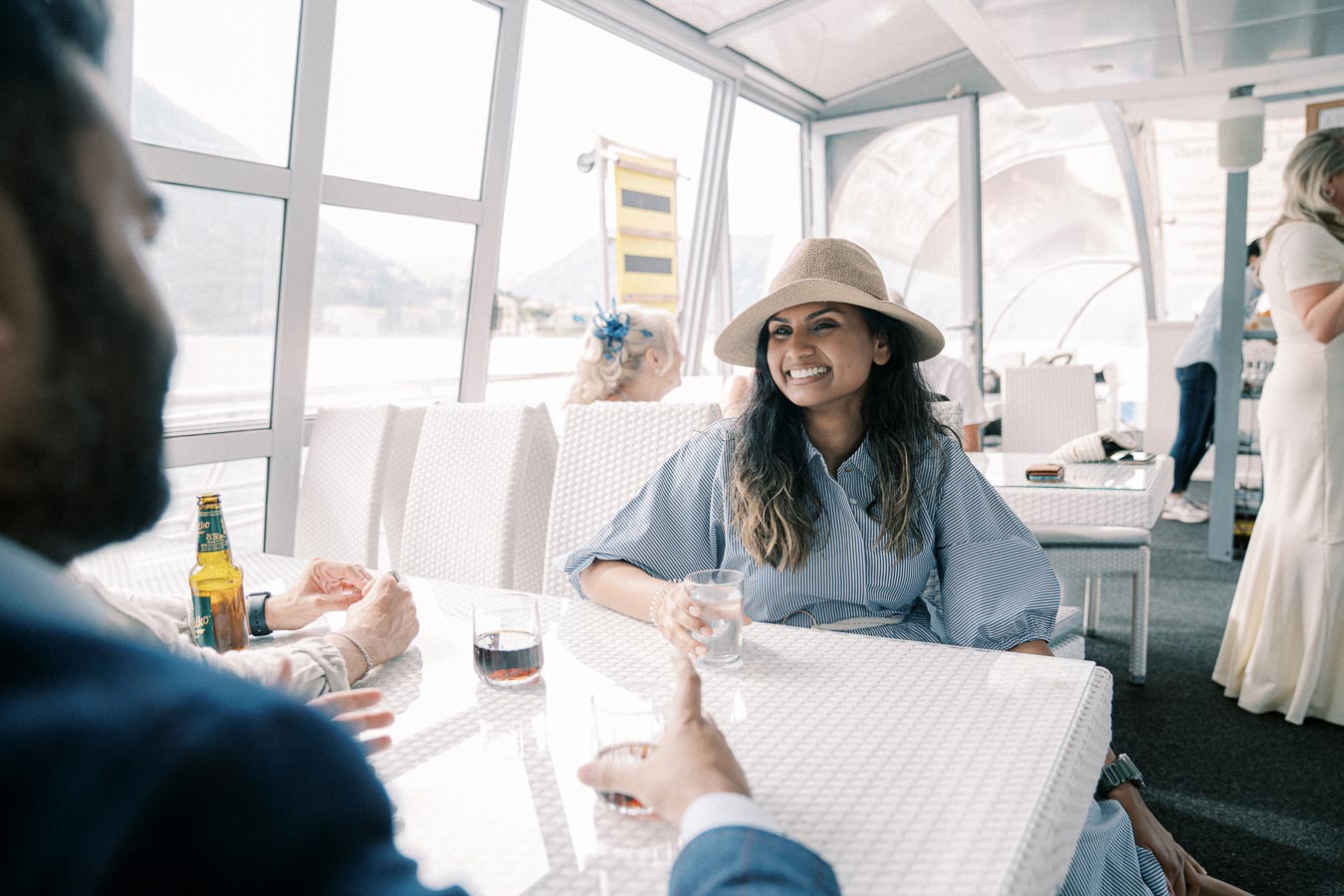 A woman in a hat smiling while seated at a table on a boat, enjoying a social gathering with drinks.
