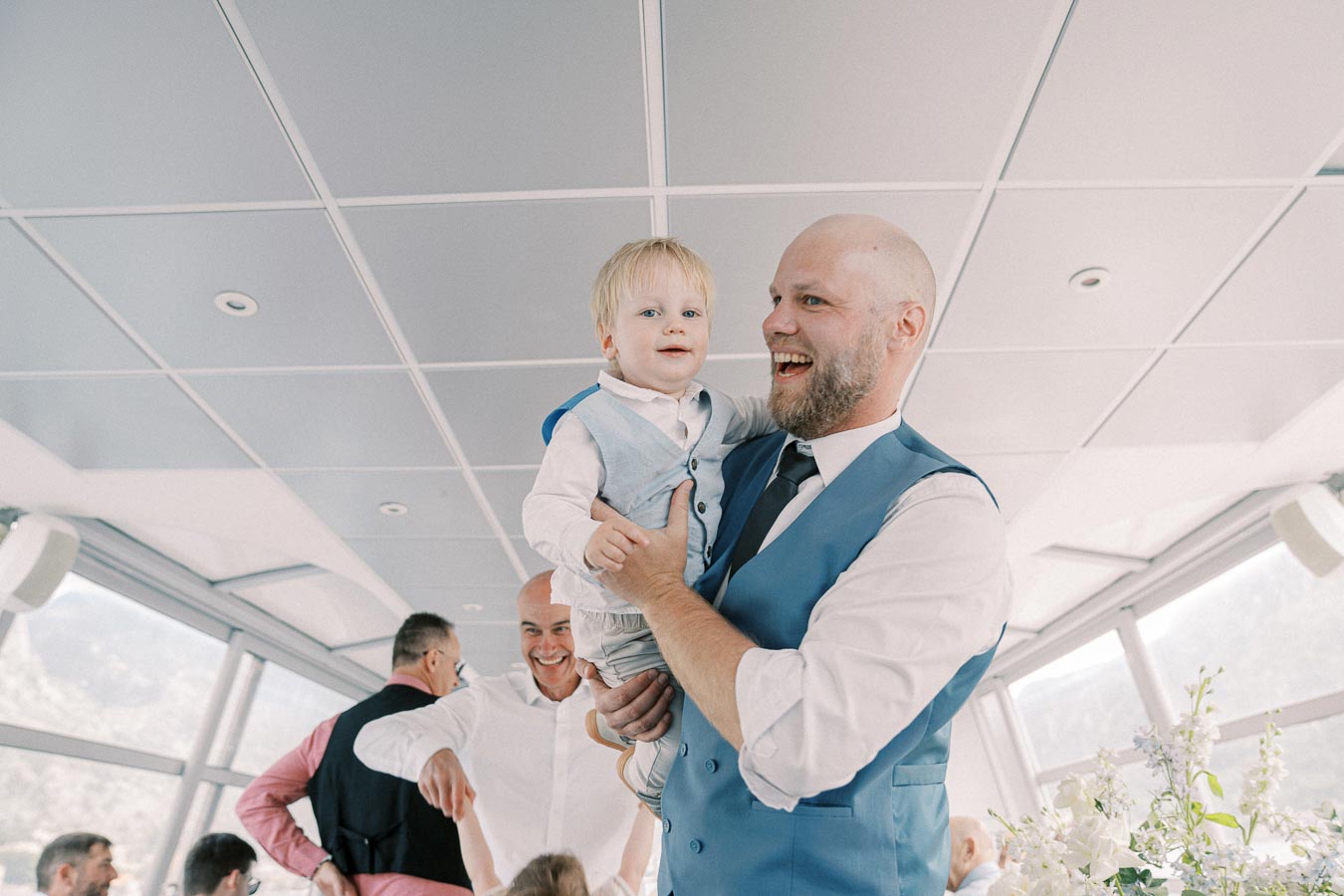 A smiling man in a blue vest holds a young child in a matching outfit at a festive indoor gathering, surrounded by people and floral arrangements.