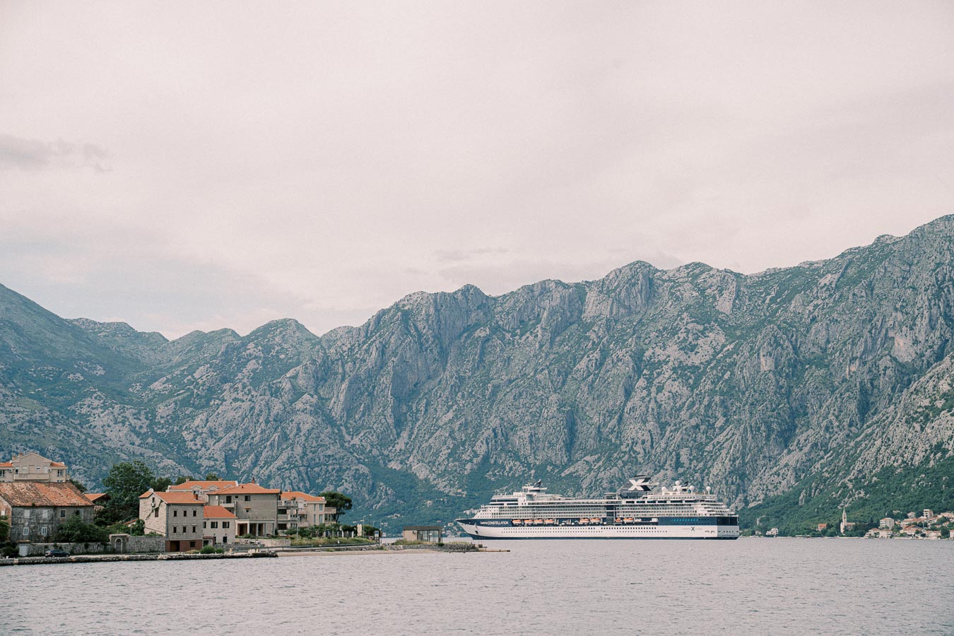Cruise ship sailing near a coastal town with mountains in the background under a cloudy sky.