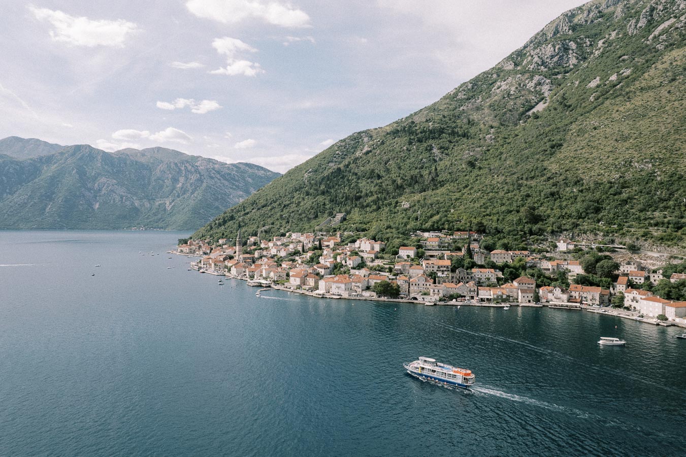 Scenic coastal view of a charming seaside town with red-roofed buildings nestled between lush green mountains and a clear blue sea, featuring a boat cruising through the water under a partly cloudy sky.