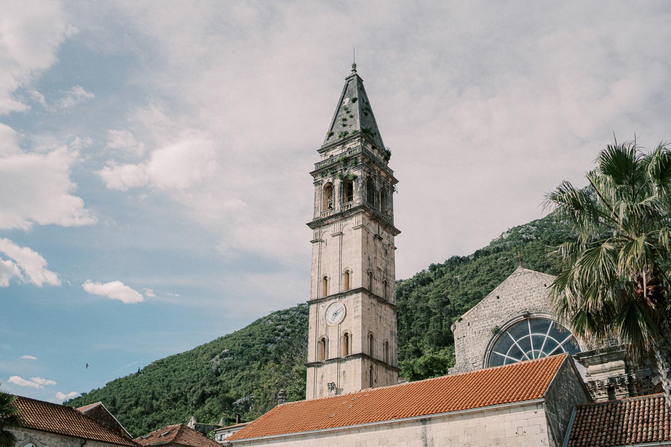 Historic stone church tower with clock against a backdrop of lush green hills and cloudy blue sky