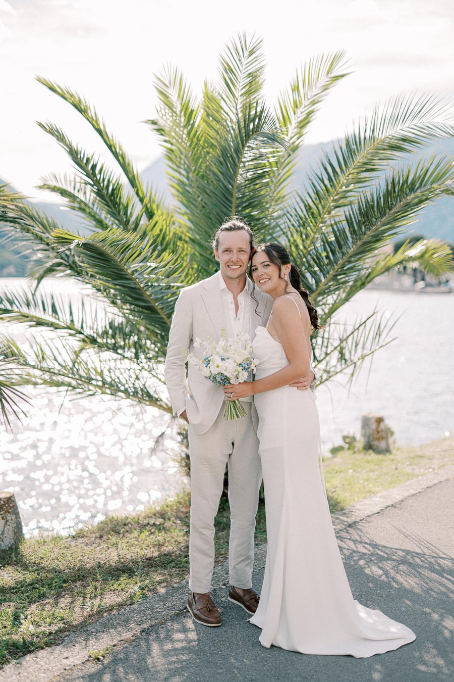 A newlywed couple poses happily by a scenic lakeside, embracing under a palm tree, with the bride in a white dress holding a bouquet of blue and white flowers, and the groom in a beige suit.