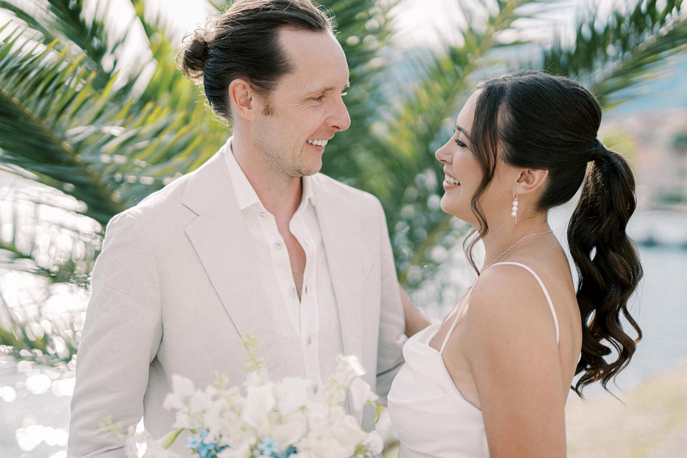 A couple in elegant attire smiling at each other in an outdoor setting with palm leaves in the background.