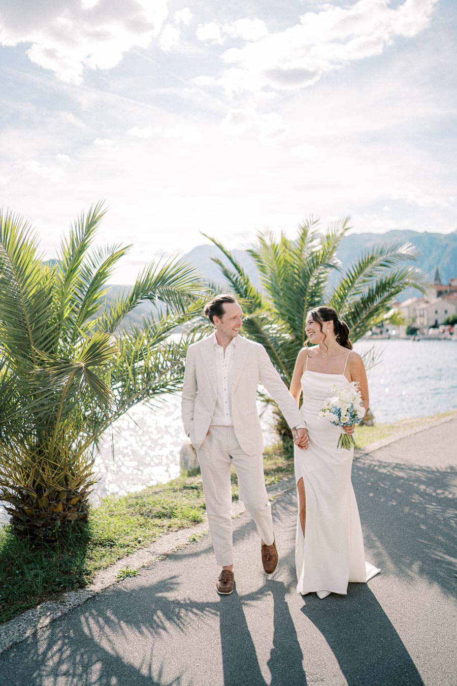 A bride and groom walking hand in hand along a scenic waterfront path lined with palm trees, under a bright, partly cloudy sky.