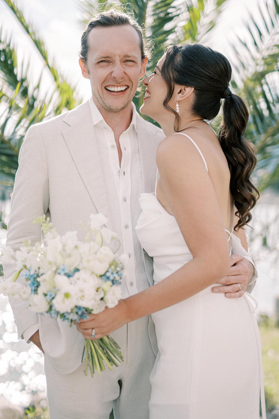 A joyful couple in elegant wedding attire embraces outdoors, with the woman holding a bouquet of white and blue flowers, set against a backdrop of palm leaves and natural light.