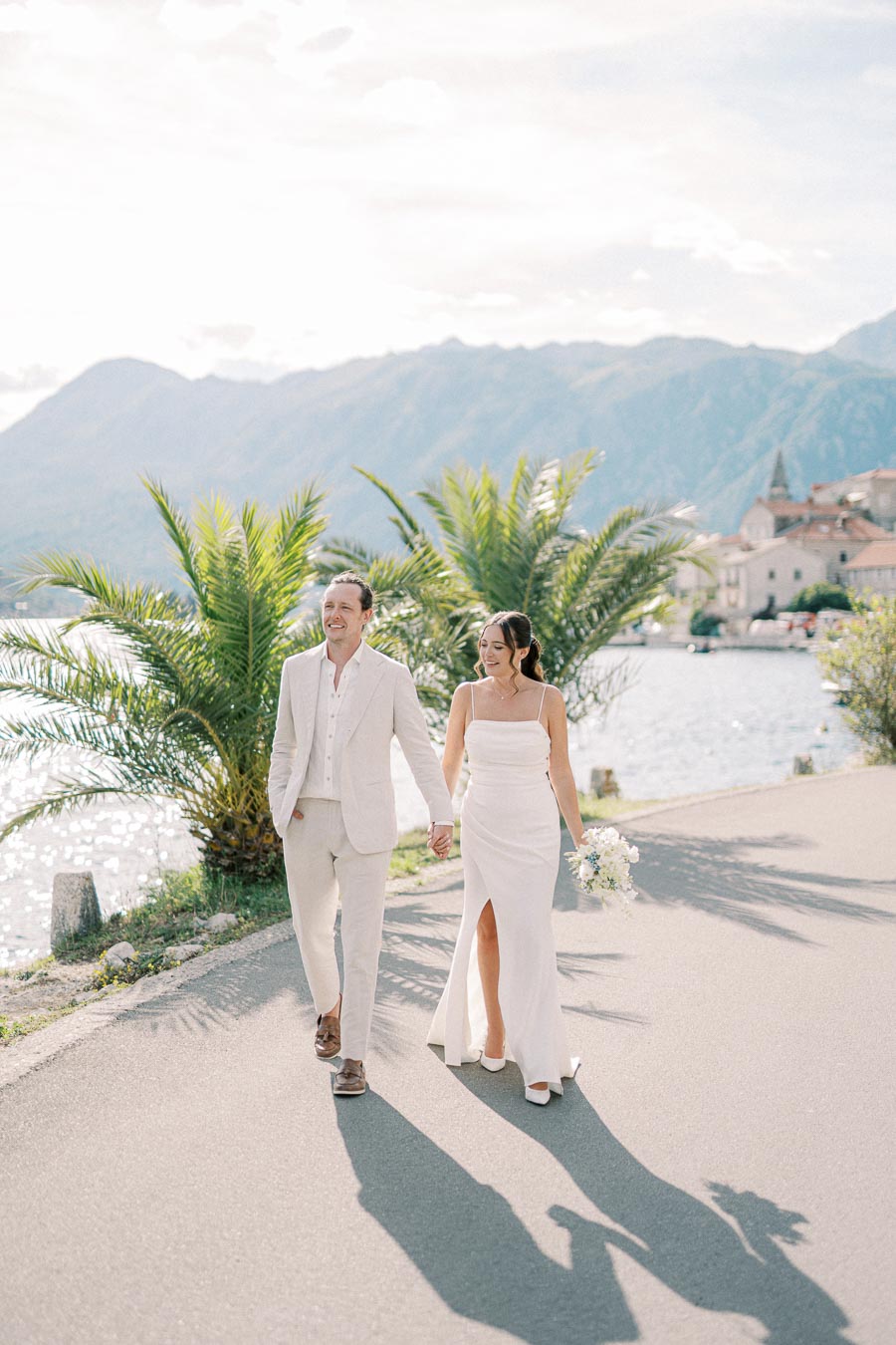 A couple in elegant white attire walks hand in hand along a scenic waterfront path, surrounded by palm trees, with a picturesque mountain landscape in the background.