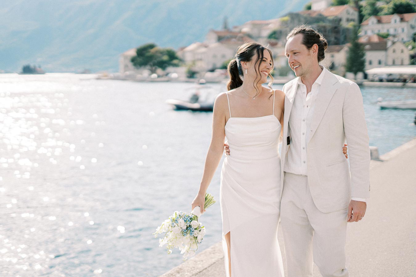 Smiling bride and groom walking by a picturesque waterfront, wearing elegant white attire, with distant mountains and quaint buildings in the background.