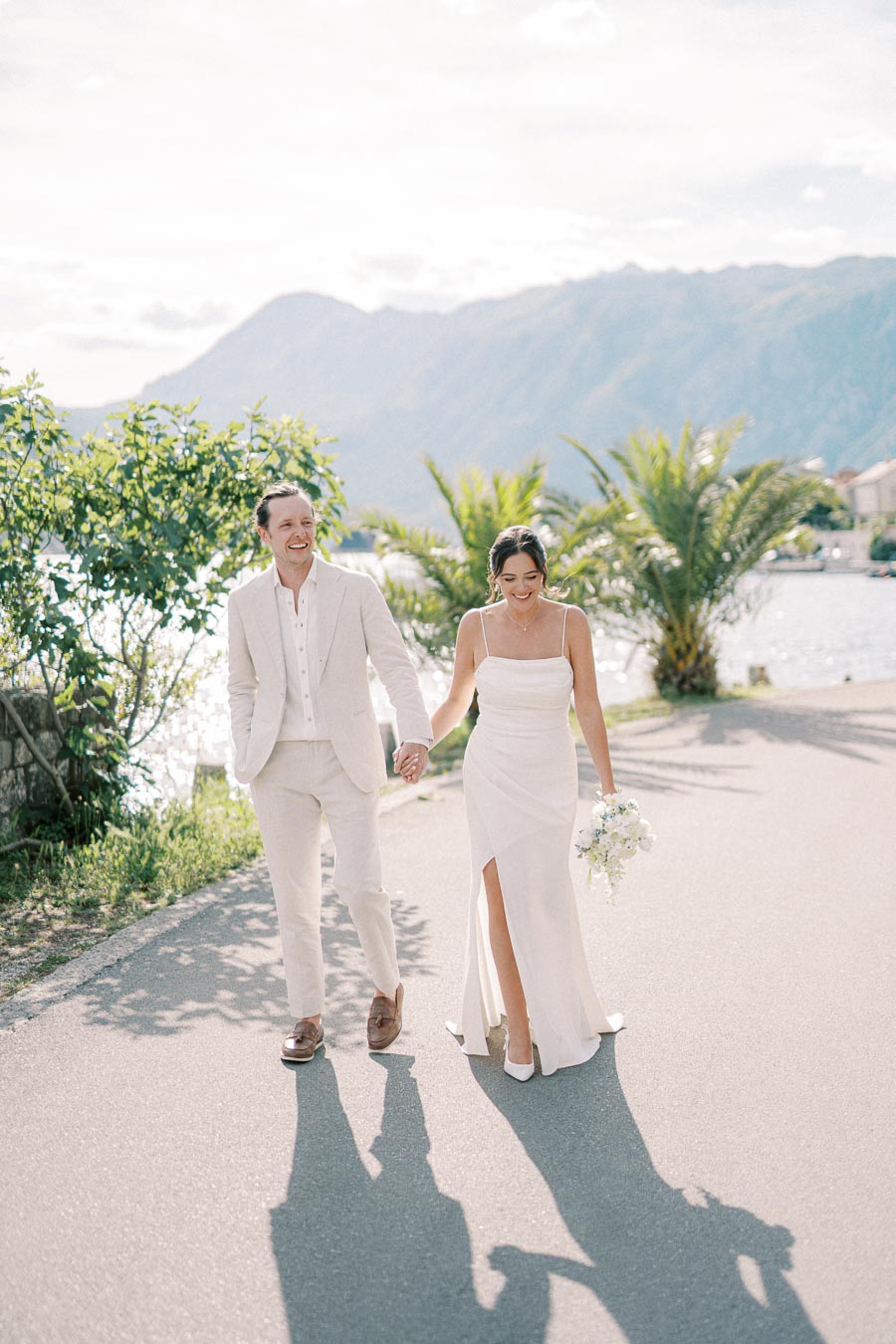 A bride and groom wearing elegant white attire walk hand in hand along a scenic waterfront path, with lush greenery and mountains in the background.