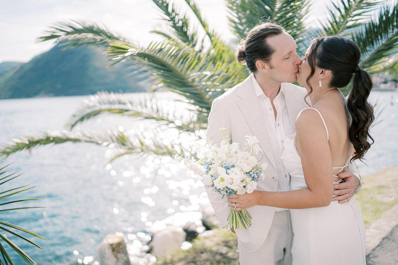 A bride and groom share a romantic kiss by a scenic lakeside, surrounded by palm trees, with the sun shining on the water. The bride holds a bouquet of white and light blue flowers, dressed in a white gown, while the groom wears a light-colored suit.