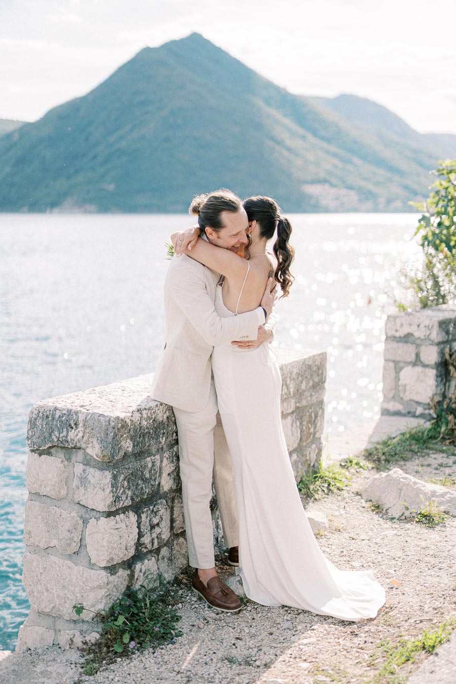 A couple in elegant wedding attire shares a romantic embrace beside a scenic waterfront with a mountain backdrop, captured in natural sunlight.