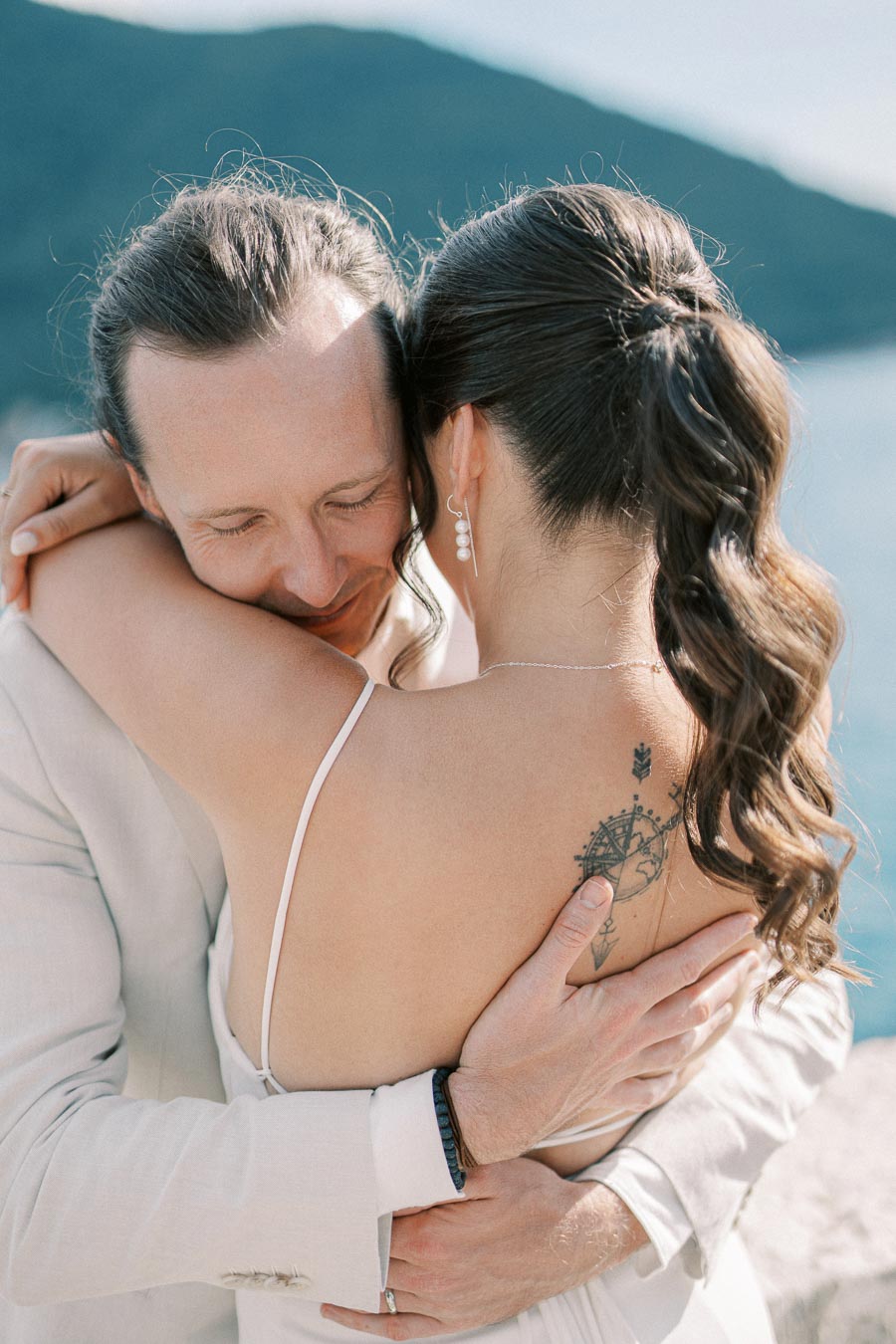 A couple embracing with joy on their wedding day, standing outdoors near a scenic water and mountain backdrop. The bride’s back displays a tattoo, and she wears elegant pearl earrings, while the groom shows a contented smile.