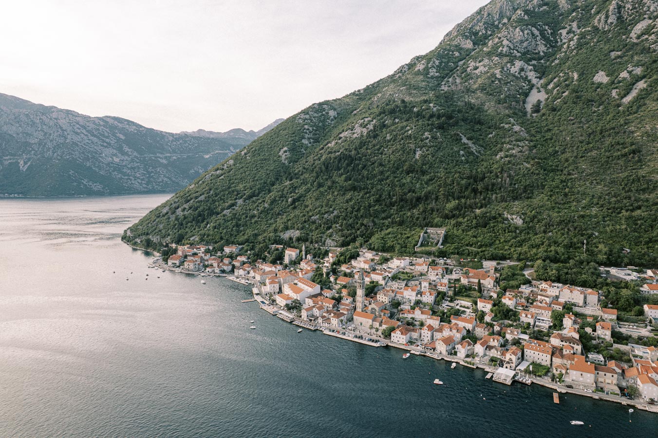 Aerial view of a picturesque coastal town with red-roofed buildings nestled at the base of a lush green mountain, adjacent to a calm blue bay on a cloudy day.