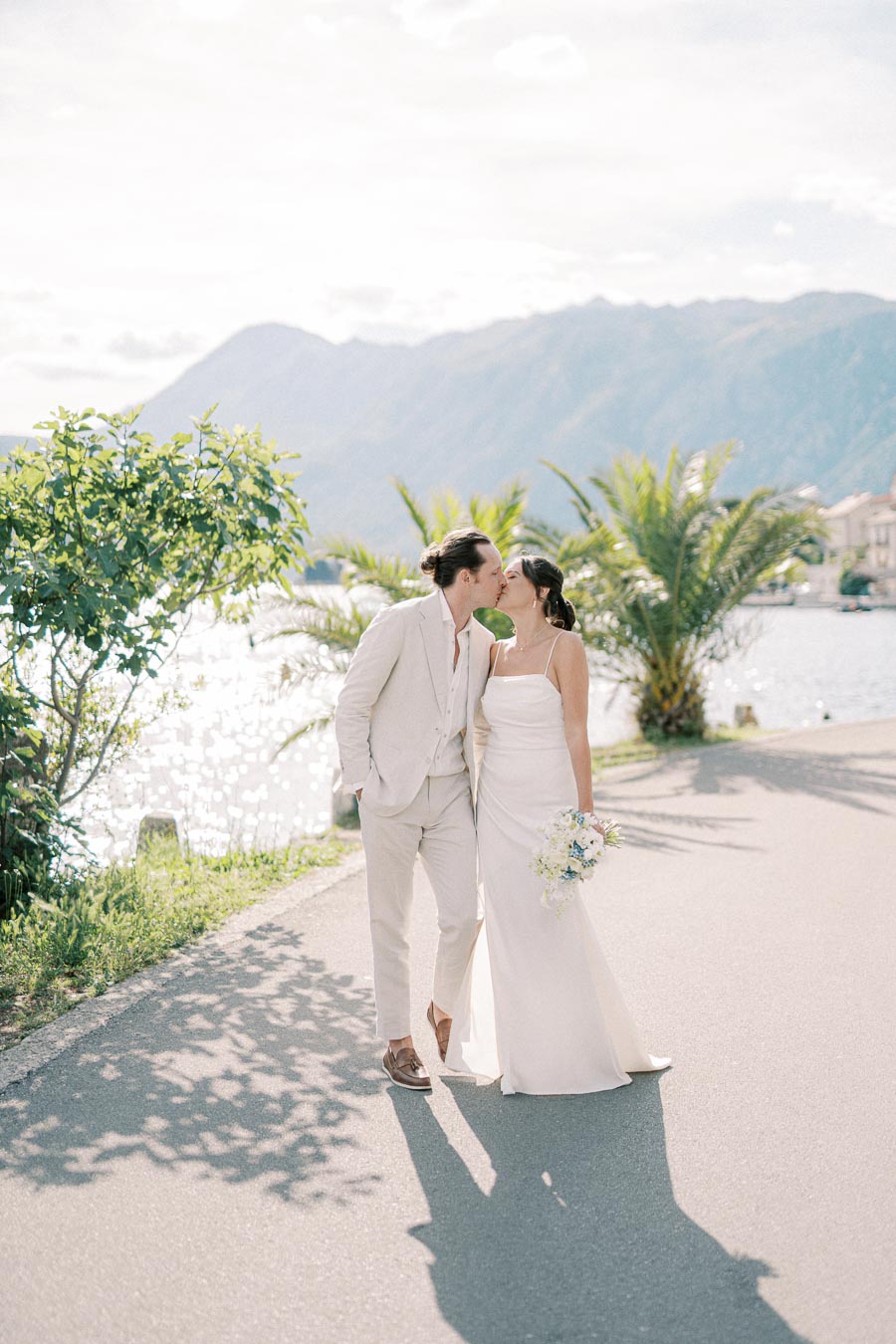 A bride and groom share a romantic kiss on a waterside path with scenic mountains in the background, surrounded by palm trees and greenery, under a bright sunny sky.