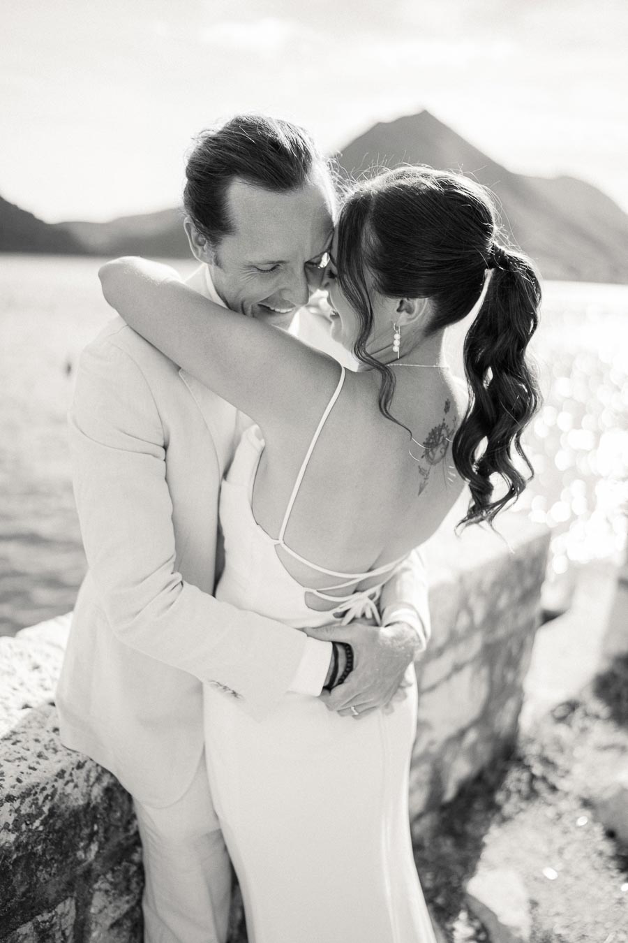 A black and white photo of a couple embracing near a scenic body of water, with a mountainous backdrop, highlighting a romantic and joyful moment.