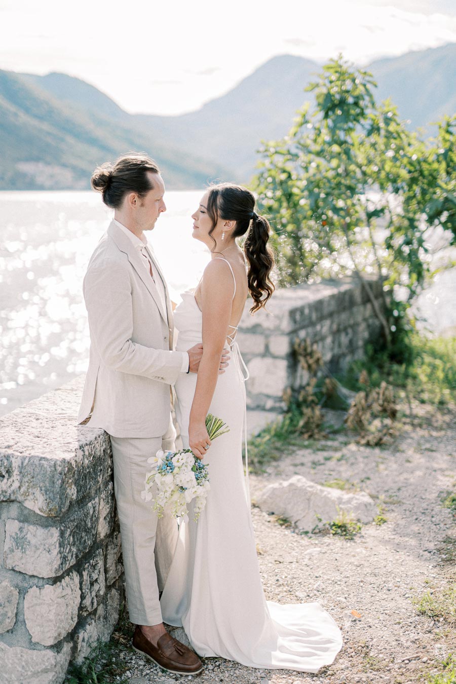A bride and groom in elegant white attire gaze into each other's eyes, standing by a stone wall with a scenic view of shimmering water and mountains in the background. The bride holds a bouquet of white and blue flowers, conveying romance and serenity in a natural setting.