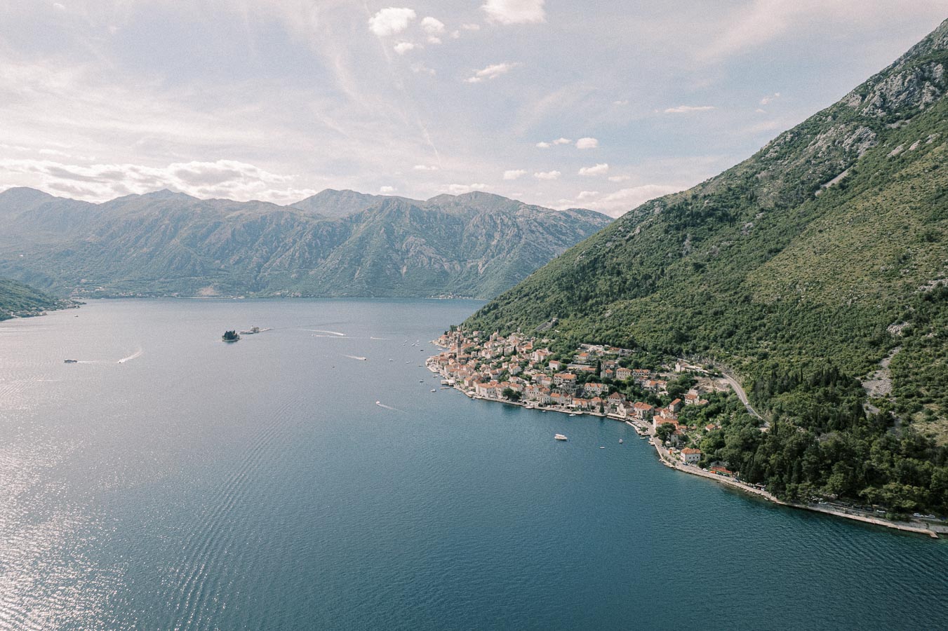 Aerial view of a coastal village nestled between a large bay and mountainous terrain, featuring clusters of traditional buildings surrounded by lush greenery and clear blue waters under a cloudy sky.