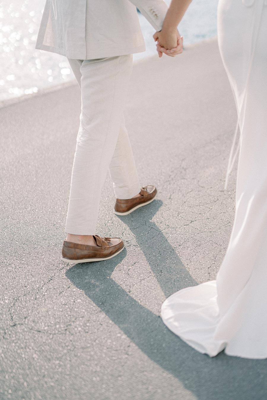 A couple wearing elegant attire walks hand in hand along a sunlit seaside path, with the bride's white dress trailing on the pavement and the groom sporting stylish brown loafers.