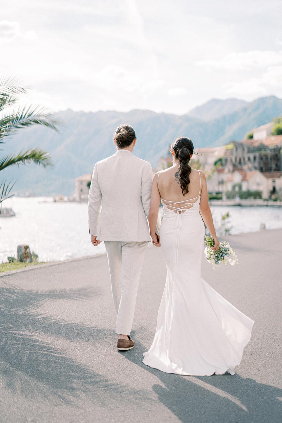 A bride and groom in elegant wedding attire walk hand-in-hand along a scenic waterfront, with mountains and charming buildings in the background under a clear blue sky.
