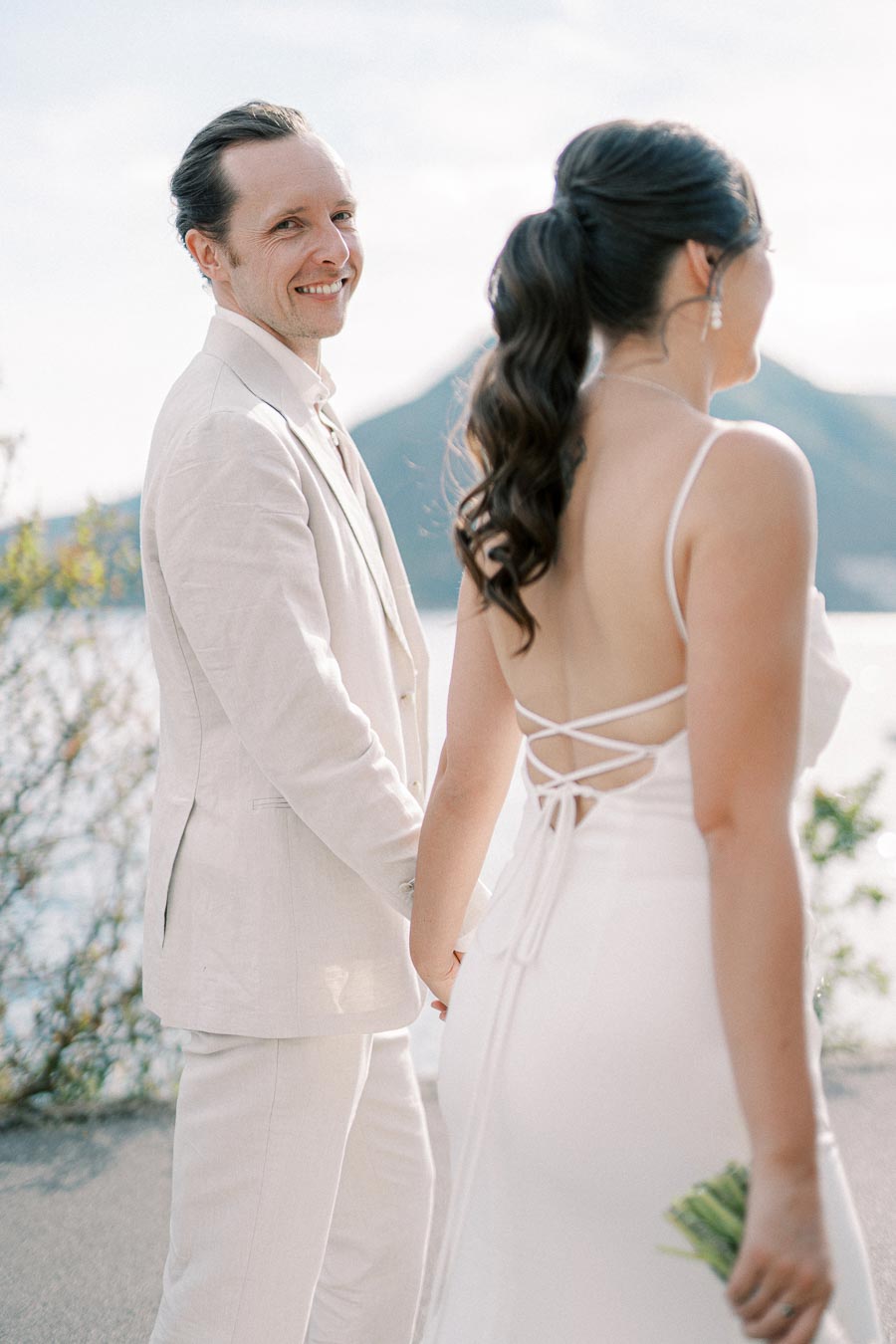 A smiling couple holding hands on their wedding day, dressed in elegant white attire, with a scenic backdrop of a lake and mountains under a clear sky.