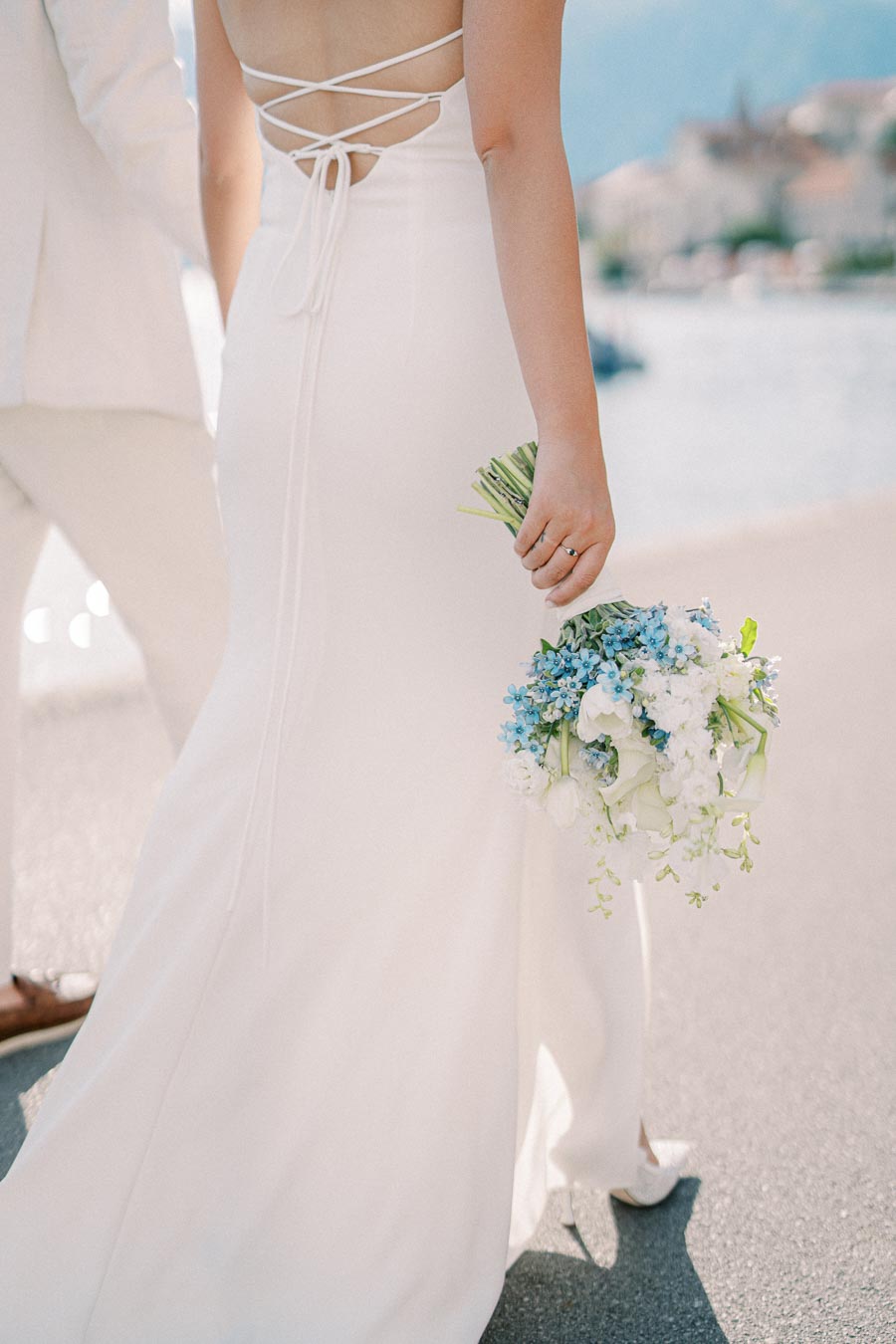 A bride in a white, backless dress holding a bouquet of white and blue flowers walks along a waterfront pathway.