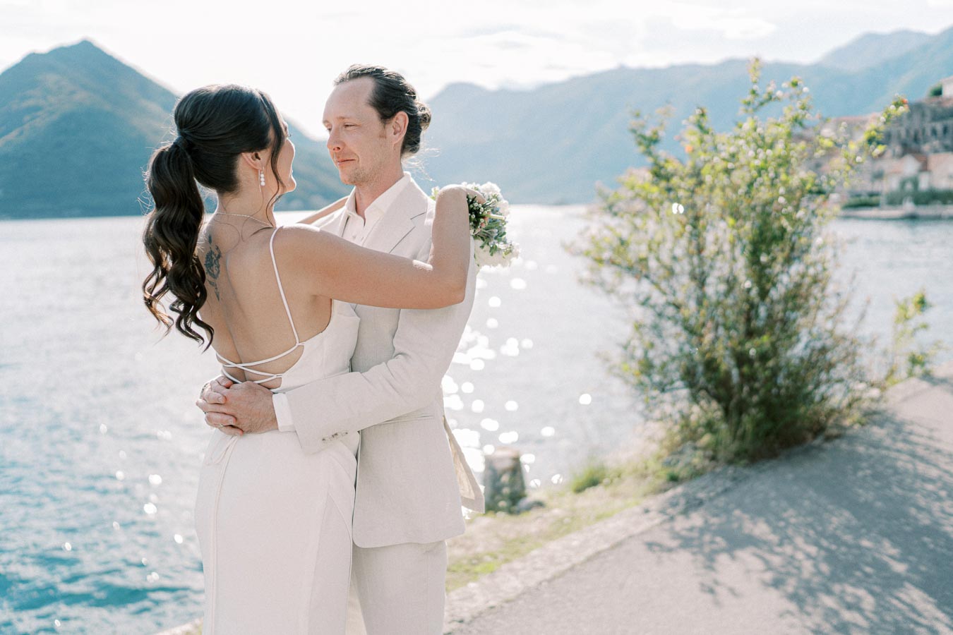 Bride and groom embracing, standing by a scenic lake with mountains in the background.