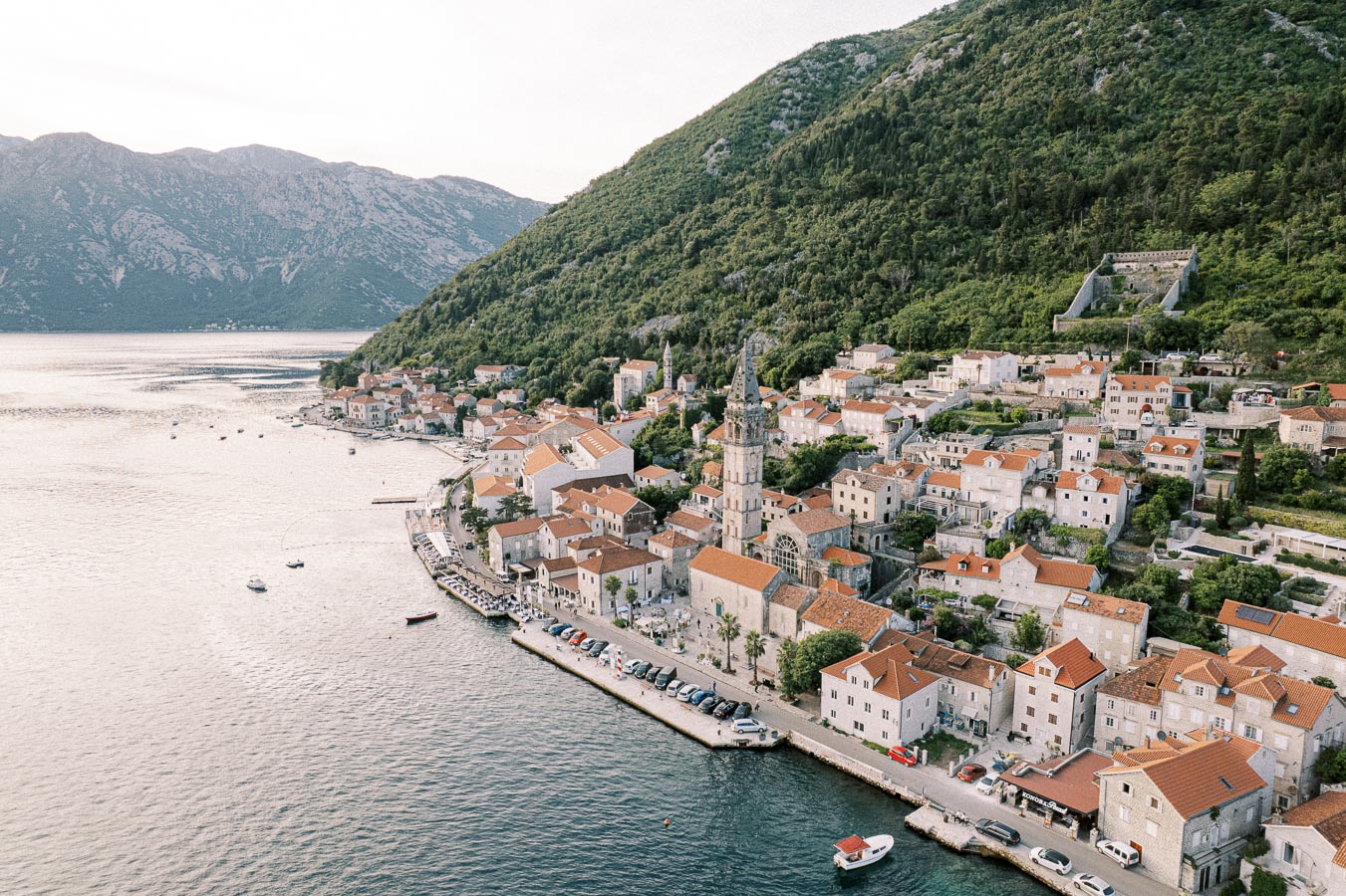 Aerial view of a picturesque coastal town with terracotta-roofed buildings nestled against green hills and overlooking a serene body of water, boats dotting the shoreline, ideal for travel and tourism content.