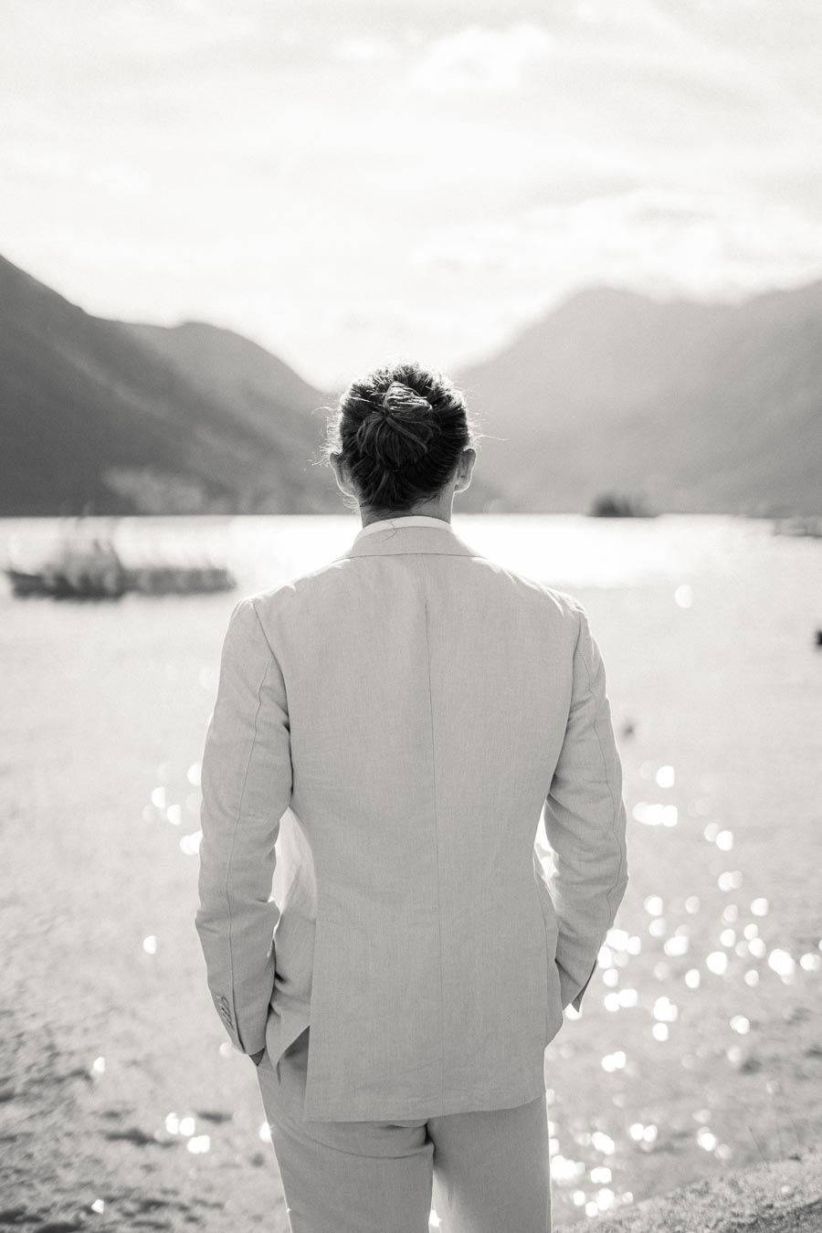 A person in a suit stands with their back to the camera, looking out over a serene lake surrounded by mountains under a bright sky.