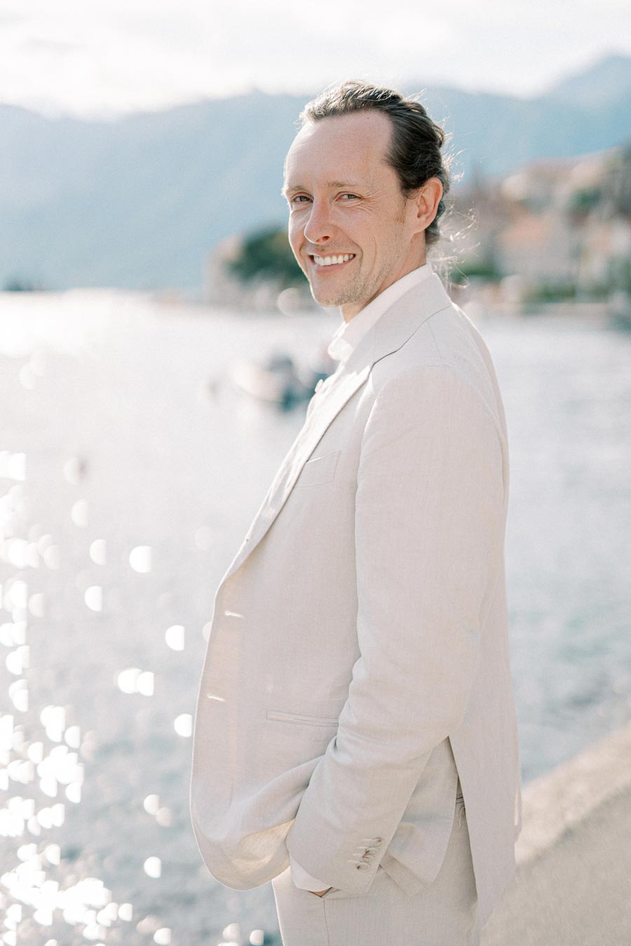 Man in beige suit smiling by a sunlit waterfront with mountains in the background.