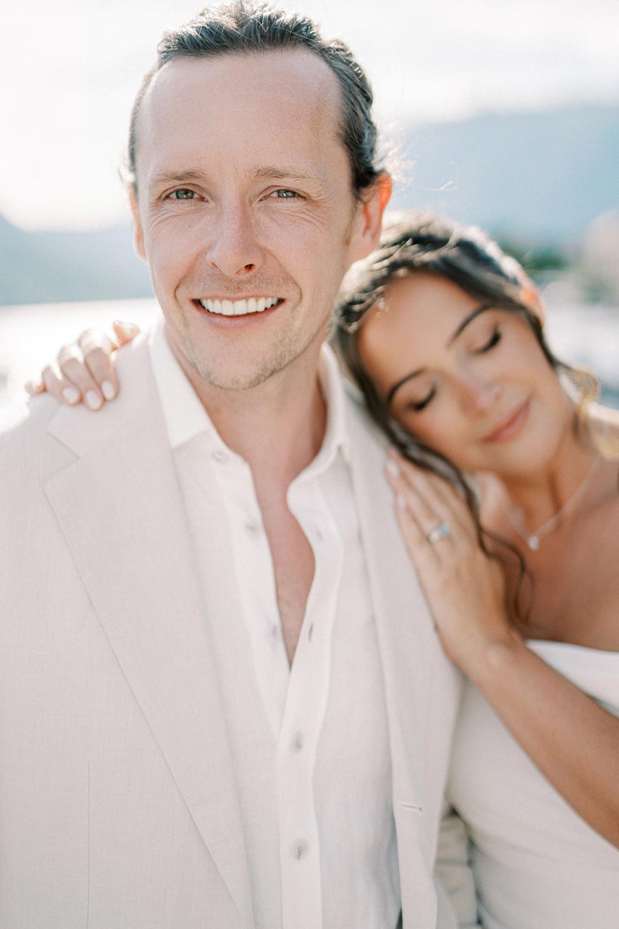 Smiling couple in elegant attire posing outdoors against a scenic backdrop, capturing a serene and romantic moment.