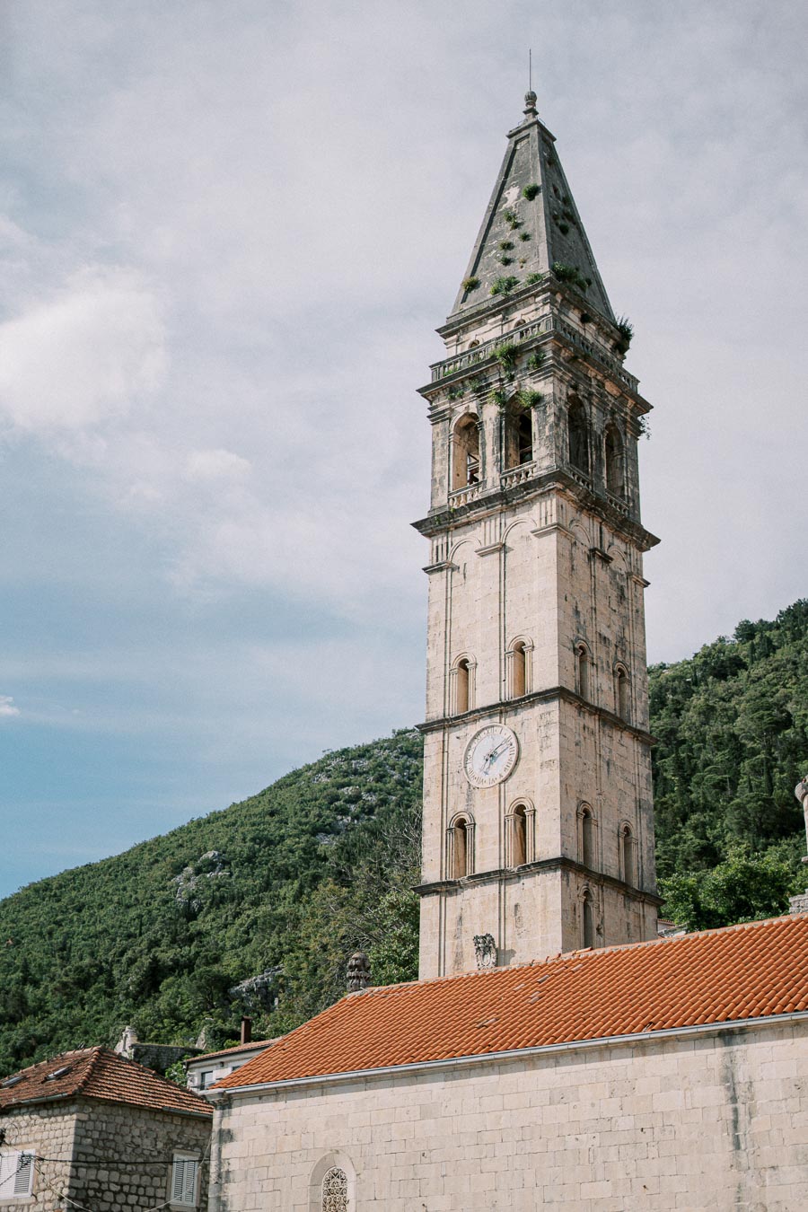 Historic stone clock tower with red-tiled roof, set against a backdrop of lush green hills and a partly cloudy sky.