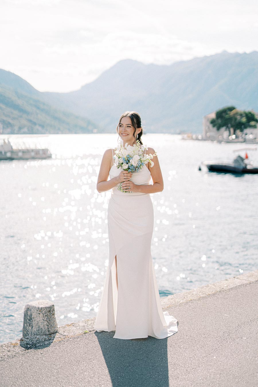 Beautiful bride in an elegant white wedding dress holding a bouquet, standing by a serene waterfront with sparkling sunlight reflecting on the water, picturesque mountains in the background.
