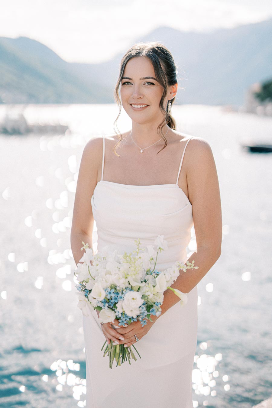 Bride in a white dress holding a bouquet of white and blue flowers by a sparkling waterfront with mountains in the background.