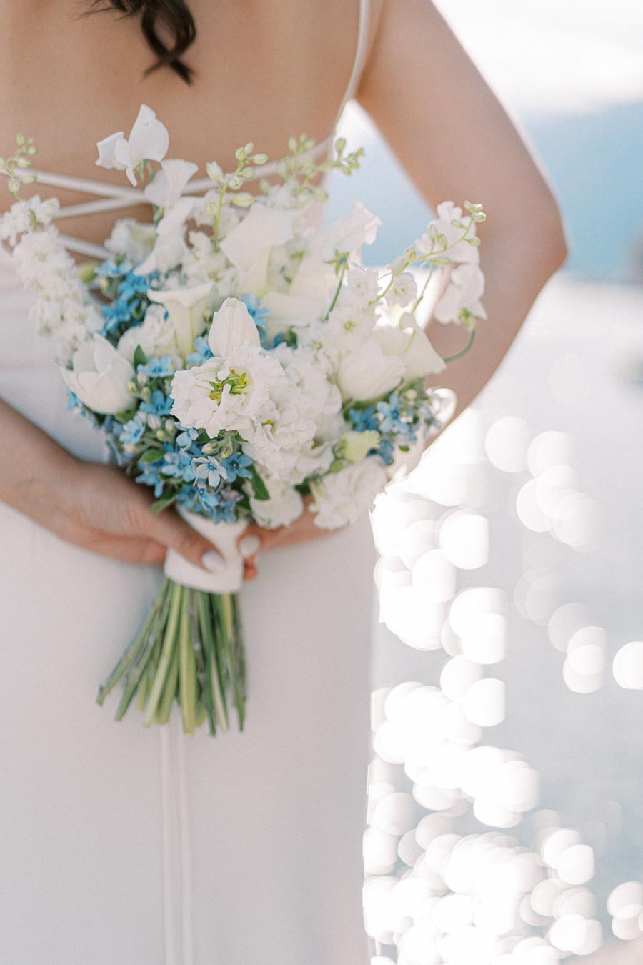 Bride holding a bouquet of white and blue flowers against a sparkling waterfront background, showcasing romantic elegance.
