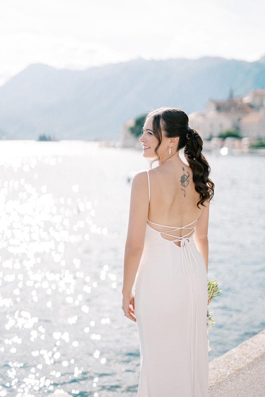 Bride in an elegant white dress with a back tattoo, standing by a serene lake with mountains in the background.