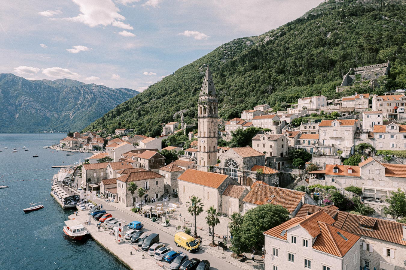 Aerial view of a coastal town with historic stone buildings and a tall church tower, nestled between lush green hills and a calm blue bay dotted with boats, under a partly cloudy sky.