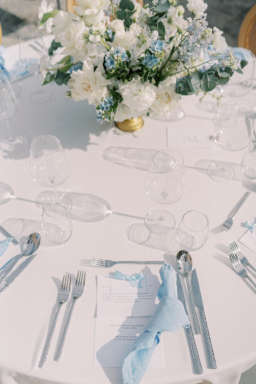 Elegant wedding table setting with a white tablecloth, featuring a floral centerpiece of white and blue flowers in a gold vase. The table is set with silver cutlery, wine glasses, and blue napkins, creating a sophisticated and romantic ambiance.
