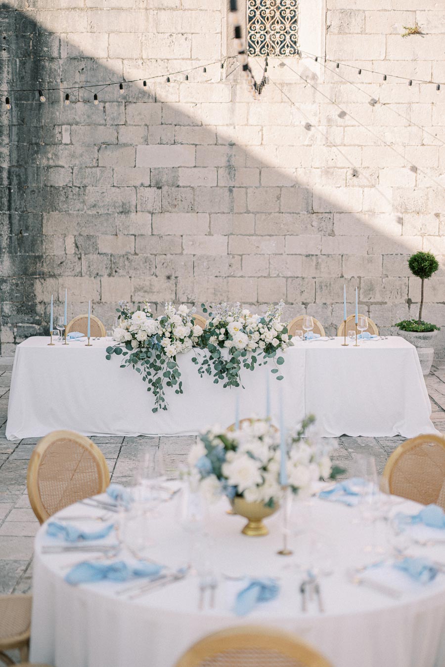 Outdoor wedding reception setup featuring elegant white table settings with blue accents, floral centerpieces, and decorative greenery against a rustic stone wall.