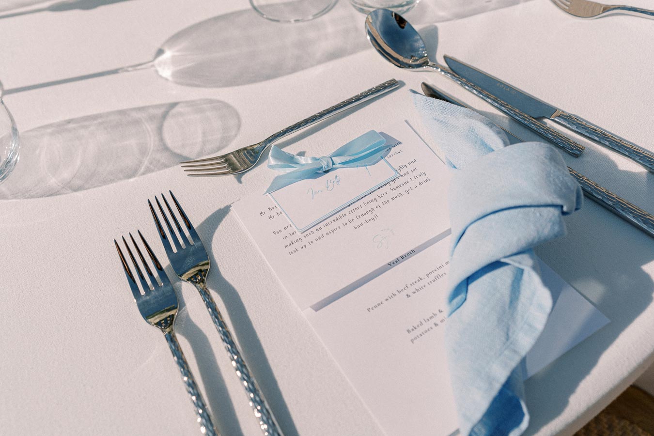 Elegant table setting with polished silverware, glasses, blue napkin, and a menu card with a blue ribbon, highlighting a refined dining experience.