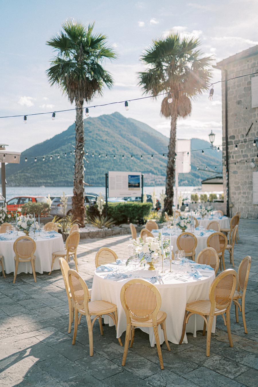 Outdoor wedding reception with elegant round tables, set with white tablecloths and blue napkins, surrounded by wooden chairs. Palm trees and mountainous landscape provide a scenic backdrop, accentuated by string lights creating a romantic atmosphere. Ideal venue for a picturesque celebration by the water.