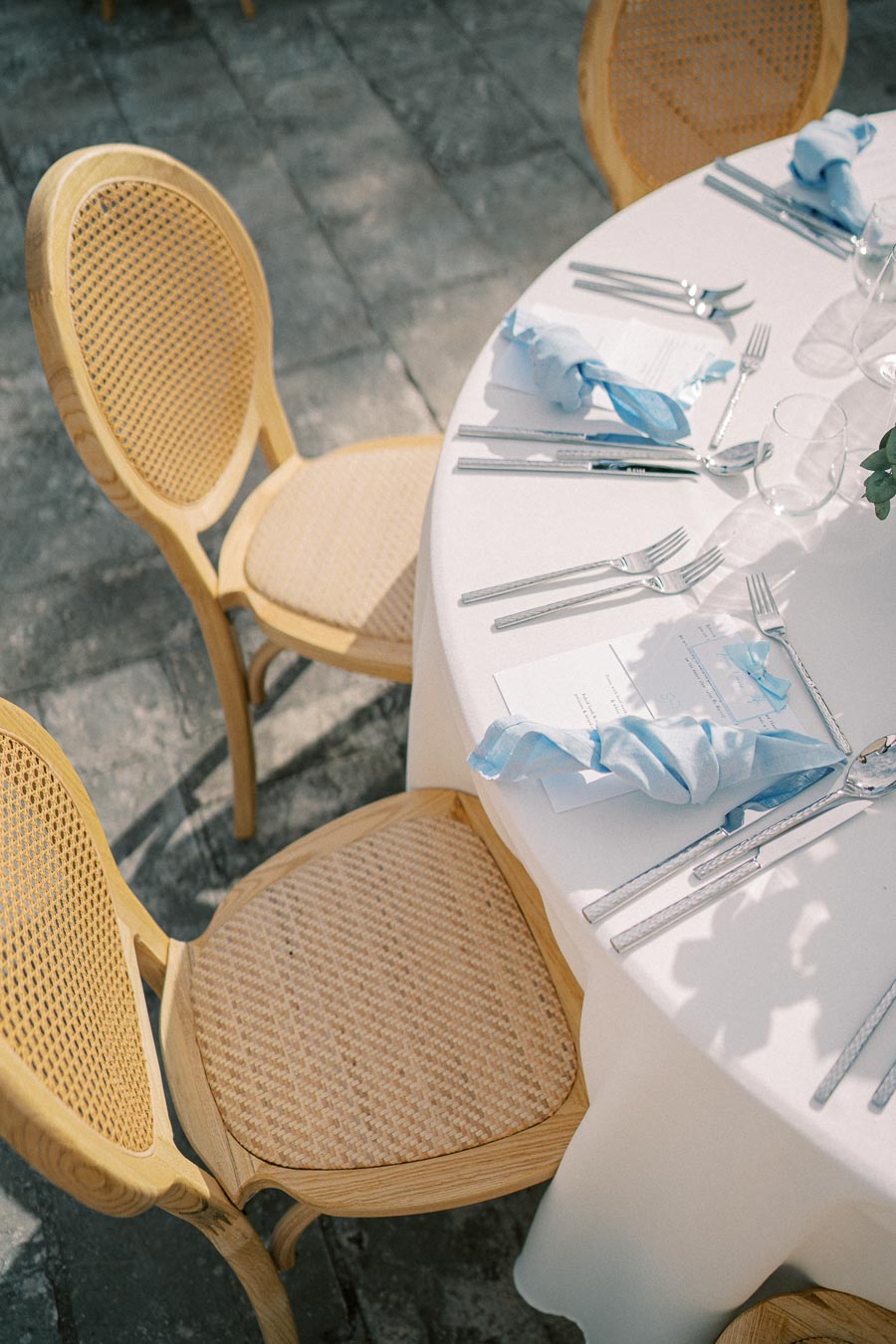 Outdoor event table setting with wicker chairs and white tablecloth; adorned with polished silverware, glassware, and blue napkins under natural light.