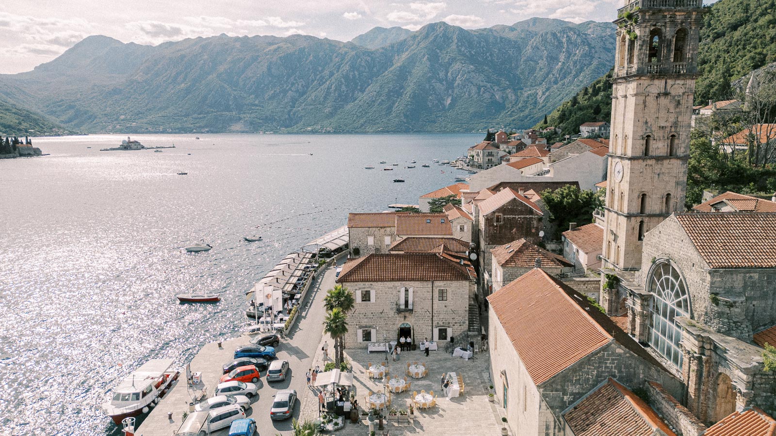 Aerial view of the scenic coastal town of Perast in Montenegro, featuring historic stone buildings with red-tiled roofs, a prominent clock tower, and the tranquil waters of Kotor Bay framed by majestic mountains in the background.