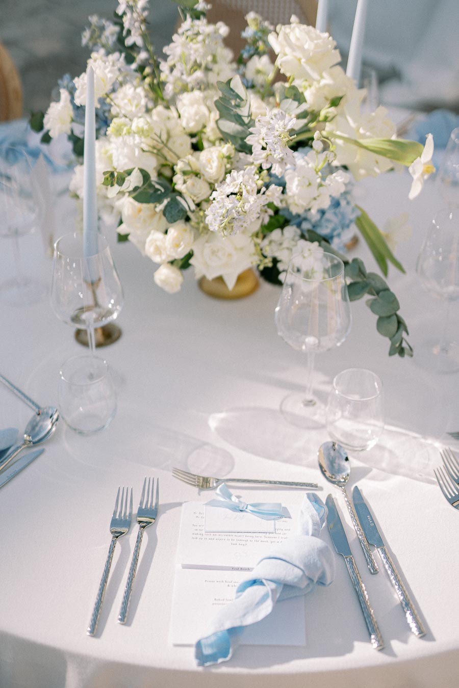 Elegant wedding table setting featuring a white tablecloth, silver cutlery, wine glasses, and a centerpiece of white and blue flowers. Blue napkins and candles add a touch of color to the sophisticated decor.